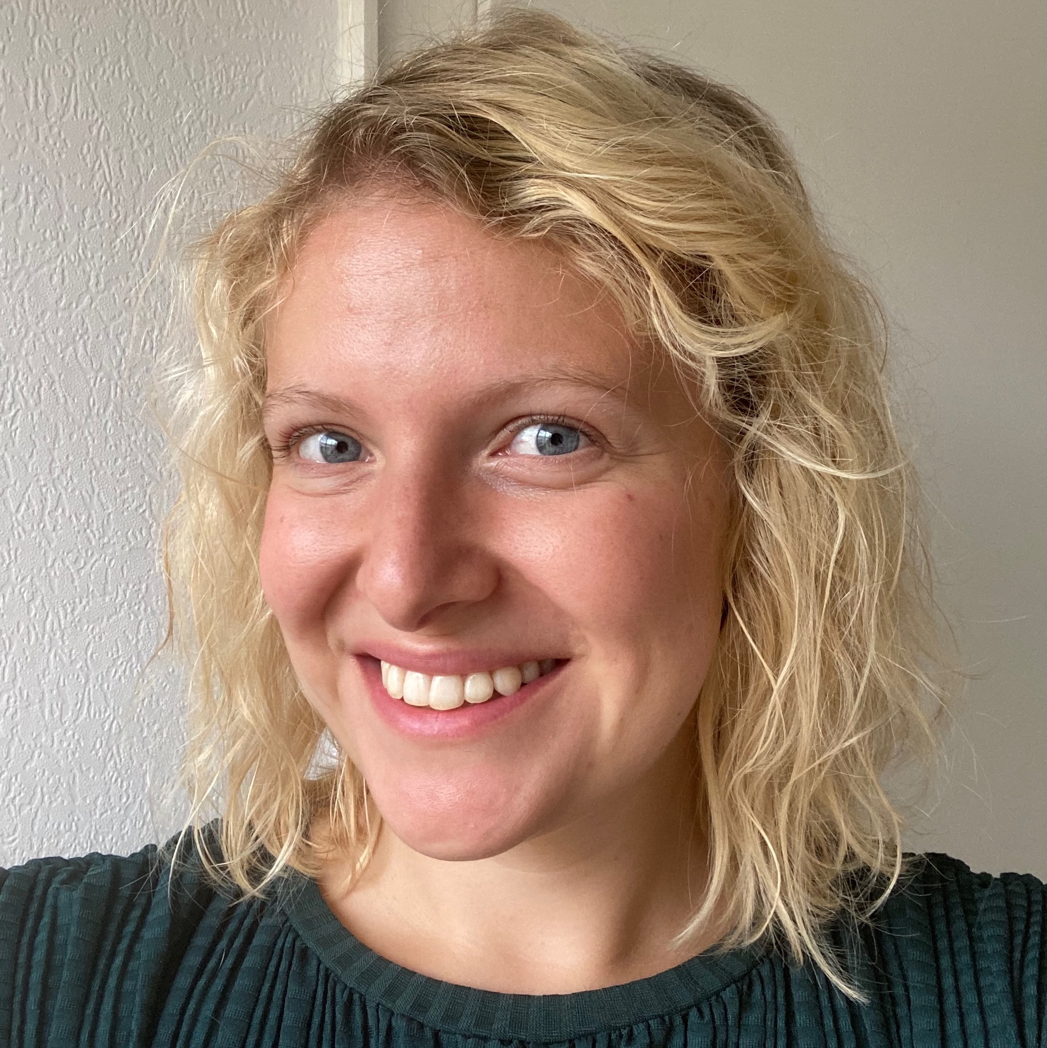 Mandy lächelt dich freundlich an. Sie trägt ein dunkelgrünes Oberteil mit Rundhalsausschnitt. Ihre hellblonden, gewellten Haare sind offen und reichen bis zu ihren Schultern. Kostümbildnerin, Kostümbild, Kostümbildassistenz, Garderobiere, Styling, Stylist, Modedeisgnerin, Modedesign, Filmschaffende, Deutschland, Filmset